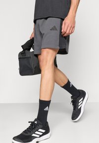 Γκρι αθλητικό σορτς με μαύρες λεπτομέρειες, σε συνδυασμό με μαύρες κάλτσες και μαύρα αθλητικά παπούτσια με λευκές ρίγες. Κρατώντας μία μαύρη τσάντα γυμναστηρίου.