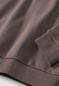 Šedo-hnědá mikina s žebrovanými manžetami a lemem, hladkou texturou a volným střihem. Detailní záběr zvýrazňuje texturu látky a švy.