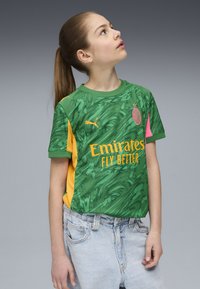 Maglia verde con motivo astratto, dettagli gialli e particolari rosa. Presenta il logo e il testo "Emirates Fly Better." Design aderente, maniche corte.