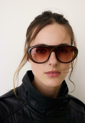 Mladá žena so svetlou pokožkou a hnedými vlasmi nosí veľké hnedé slnečné okuliare a čiernu koženú bundu na jednoduchom pozadí.