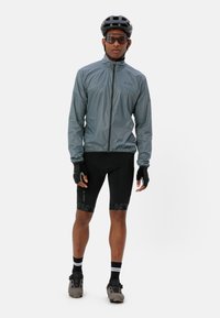 Leichte graue Radjacke mit Reißverschluss, schwarze gepolsterte Shorts mit kontrastierendem schwarzem Muster, fingerlose Handschuhe und ein schwarzer Helm.