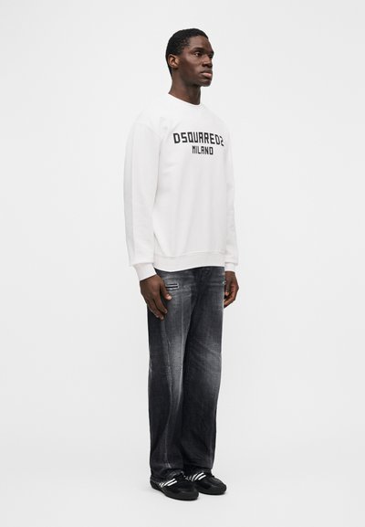 Άνδρας που στέκεται στραμμένος προς τα δεξιά φορώντας λευκή φούτερ DSQUARED2 Milano, ξεθωριασμένο μαύρο τζιν και μαύρα αθλητικά παπούτσια με λευκές ρίγες.