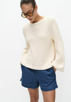 Vrouw draagt een crèmekleurige gebreide trui en blauwe denim shorts met handen in zakken, kijkend naar beneden, tegen een witte achtergrond.