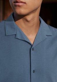 Chemise à manches courtes bleu clair avec col, motif texturé, deux boutons et encolure décontractée. Le matériau semble respirant.