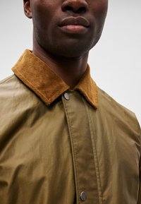 Ελαιόχρωμο σακάκι με κολάρο από καμπαρντίνα. Διαθέτει κλείσιμο με κουμπιά snap και λεία υφή, τονίζοντας έναν σύγχρονο σχεδιασμό.