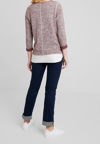 Suéter de punto color marrón burdeos con mangas tres cuartos y puños enrollados, sobre camisa blanca, combinado con vaqueros azul oscuro y zapatillas blancas.