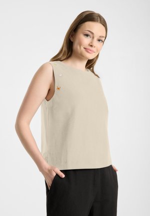 Jonge vrouw met lichtbruin haar draagt een mouwloze beige top met kleine geborduurde ontwerpen, zwarte broek, handen in de zakken, licht glimlachend.