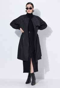 Chaqueta bomber negra con cremallera frontal, puños acanalados y un bolsillo en el pecho, combinada con una falda midi negra y botas de tacón con cordones.