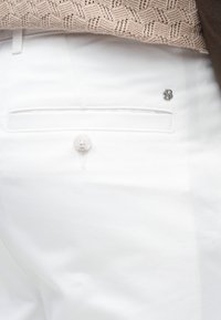 Κοντινό πλάνο της πίσω τσέπης λευκών παντελονιών με κουμπί και μικρό μεταλλικό λογότυπο, σε συνδυασμό με μπεζ πλεκτό τοπ.