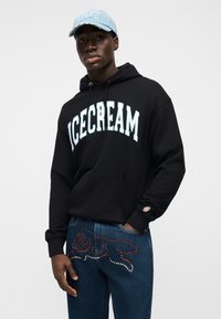 Μαύρο φούτερ με κουκούλα με τη λέξη "ICECREAM" σε ανοιχτό μπλε γράμματα· σκούρο τζιν με σχέδιο από κόκκινα και ασημένια στρας. Ανοιχτό μπλε καπέλο.