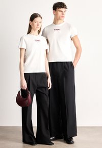 Άσπρα βαμβακερά t-shirts με το κόκκινο λογότυπο "TOMMY", σε συνδυασμό με μαύρο παντελόνι με φαρδιά παντελονοποδιά. Η γυναίκα κρατά μια μικρή μπορντό τσάντα.