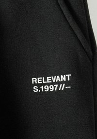 Schwarzer Sweatshirtstoff mit weißem Text: "RELEVANT S.1997 // --" auf der Vorderseite gestickt, glatte Textur, keine weiteren auffälligen Designelemente.