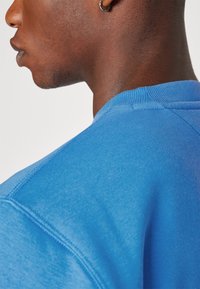 Sudadera azul con cuello redondo y textura suave, con cuello y costuras de hombro acanaladas. Primer plano de la vista trasera y lateral.