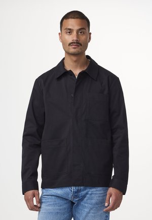 Giacca nera in tela con colletto, chiusura frontale con bottoni e due tasche sul petto. Il modello indossa jeans in denim azzurro chiaro.