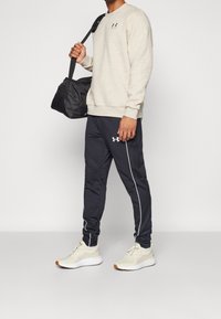 Man som bär beige sweatshirt, svarta träningsbyxor med vita ränder, krämfärgade sneakers och håller en svart gymväska över axeln.