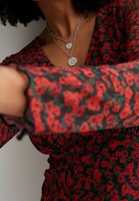 Rote Bluse mit floralen Mustern aus transparenter schwarzer Stoff, mit V-Ausschnitt und rüschengesäumten Ärmeln. Zwei geschichtete silberne Halsketten mit runden Anhängern.