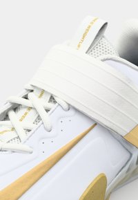 Zapatilla deportiva blanca con un upper texturizado, acentos dorados, una correa ancha en la parte media del pie y un collar acolchado para mayor estabilidad.