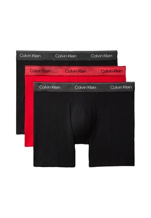 Skjorteløs mand, der står barfodet i sorte Calvin Klein boxershorts mod en ensfarvet grå baggrund, med den ene hånd bag hovedet.