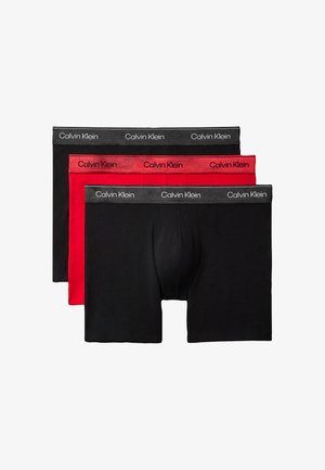 Hombre sin camiseta, de pie descalzo, con calzoncillos tipo bóxer negros de Calvin Klein sobre un fondo gris liso, con una mano detrás de la cabeza.
