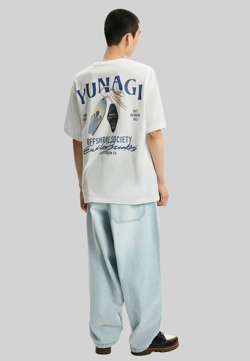 White cotton t-shirt featuring a graphic print on the back, paired with light blue baggy denim pants and black shoes with white accents.