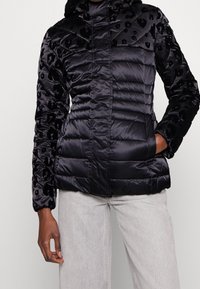 Doudoune noire avec une texture brillante en imprimé léopard sur les épaules et les manches, dotée d'une fermeture éclair et d'une capuche. Matériau léger.