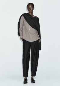 Long-sleeve, beige linen top paired with black, loose-fitting trousers. A dark, textured wrap is draped over one shoulder. Black shoes complete the look.
