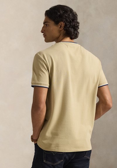 Μπεζ κοντομάνικη μπλούζα με απλό σχέδιο, σκούρο μπλε περιγράμματα στο γιακά και τα μανίκια, κατασκευασμένη από απαλό, υφασμένο ύφασμα.