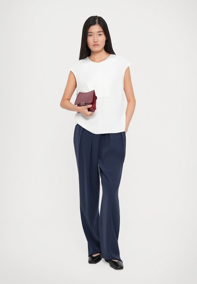 Woman with long dark hair wearing a white sleeveless top, navy wide-leg trousers, black shoes, and holding a small red handbag.