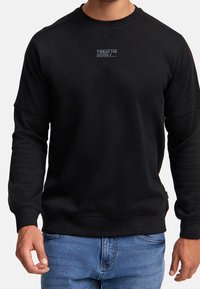 Sort sweatshirt med lille hvid tekst "WEAR THE CODE" på brystet, båret af en person i blå jeans, der står imod en ensfarvet baggrund.