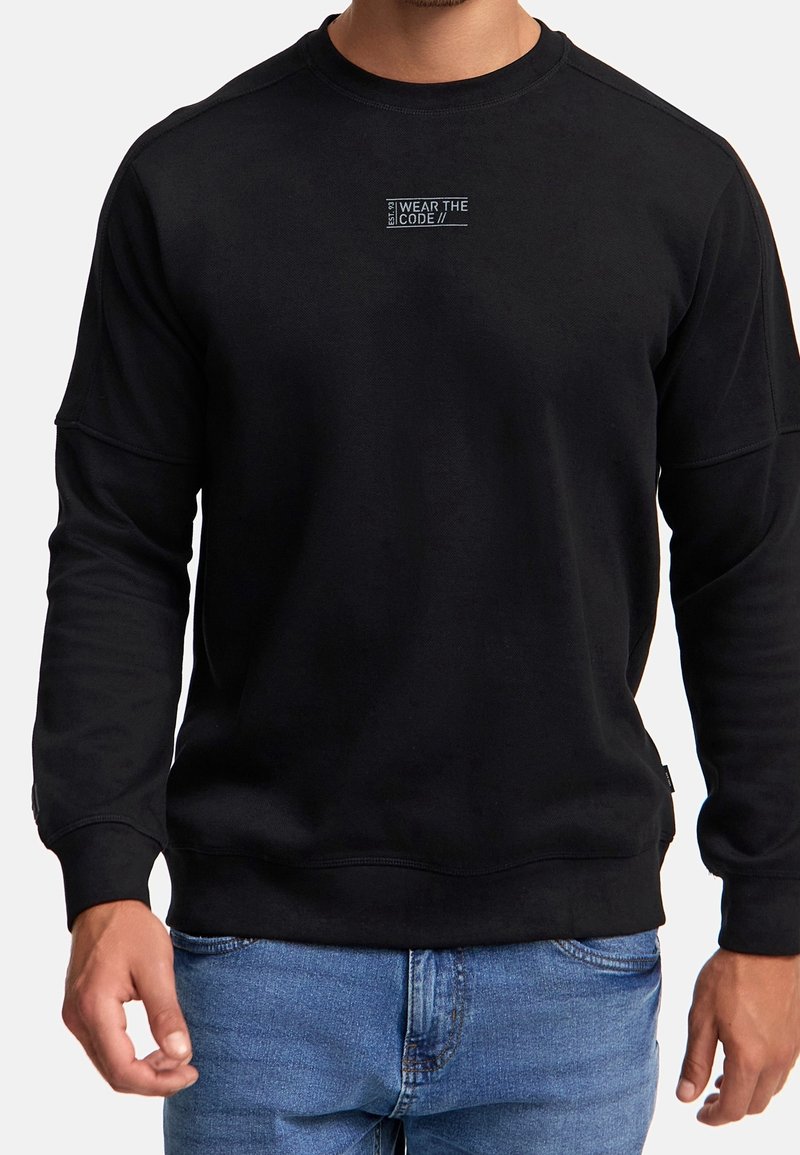 Sort sweatshirt med lille hvid tekst "WEAR THE CODE" på brystet, båret af en person i blå jeans, der står imod en ensfarvet baggrund.