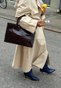 Borsa a tracolla in pelle marrone con stampa coccodrillo; trench coat beige; stivaletti blu testurizzati con punta affilata e tacchi a stiletto; in mano limone e bottiglia.