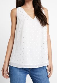 Vit ärmlös topp med V-hals, prydd med metalliska silverprickar, något genomskinligt tyg och lös passform, kombinerad med blå jeans.