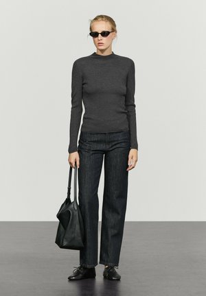 Femme portant des lunettes de soleil noires, un pull ajusté gris foncé, un jean noir, tenant un grand sac à main en cuir noir, debout devant un fond uni.
