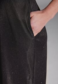 Tejido negro con un sutil brillo, que cuenta con un bolsillo lateral y una textura suave. El tejido cae con elegancia, destacando su diseño a medida.