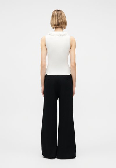 Άτομο με κοντά ξανθά μαλλιά, φορώντας ένα λευκό αμάνικο τοπ με βολάν στη λαιμόκοψη και φαρδιά μαύρη παντελόνα, στέκεται γυρισμένο με την πλάτη.