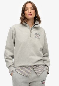 Sudadera gris claro con cremallera hasta el cuarto, con la impresión "SUPERDRY ATHLETICS", que presenta una textura suave, un corte relajado y una camisa de rayas en capas debajo.