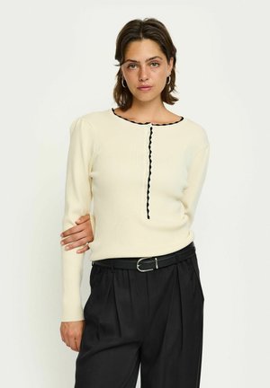 Pull crème côtelé avec une bordure noire en forme de coquille au niveau du col et un détail de boutons, associé à un pantalon noir taille haute et une ceinture noire.