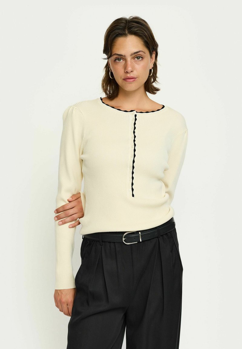 Gerippter cremfarbener Pullover mit schwarzem gewelltem Saum am Ausschnitt und Knöpfen, kombiniert mit hochgeschnittenen schwarzen Hosen und einem schwarzen Gürtel.
