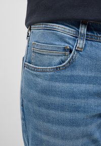 Primer plano del bolsillo y la trabilla del cinturón de unos vaqueros de mezclilla azul con costuras visibles, usados con una camisa oscura.