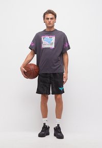 Νέος άνδρας με μαύρο σορτς μπάσκετ και γραφικό t-shirt κρατώντας μια μπάλα μπάσκετ, στέκεται μπροστά σε απλό λευκό φόντο.