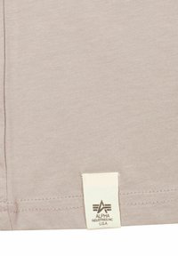 Tessuto beige con un orlo cucito e un'etichetta rettangolare bianca con la scritta "Alpha Industries Inc U.S.A" attaccata vicino al bordo inferiore.