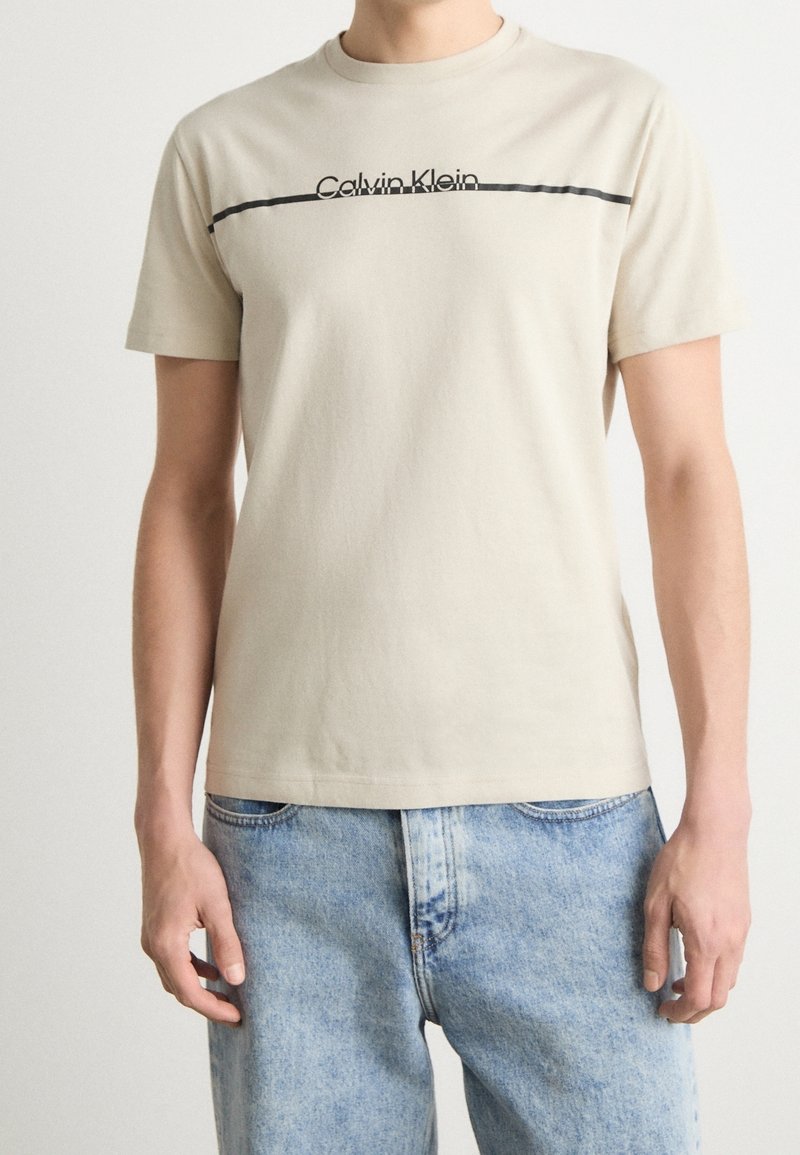 Homme portant un t-shirt beige Calvin Klein avec une ligne horizontale noire et un jean bleu, debout devant un fond blanc uni.