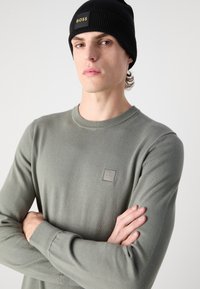 Grijze gebreide trui met een ronde hals en een klein vierkant logo-label. Het model draagt een zwarte geribde beanie. Soepele textuur en aansluitend ontwerp.