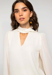 Blouse blanche avec un col en V et un détail d'écharpe drapée. Manches longues avec épaules froncées. Accessoires comprenant de grandes boucles d'oreilles en forme de goutte.