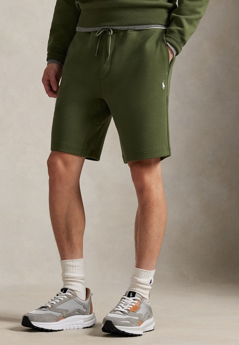 Βερμούδα από βαμβάκι σε ελιά πράσι νη απόχρωση με κορδόνι στη μέση και πλαϊνές τσέπες, συνδυασμένη με γκρι αθλητικά παπούτσια και λευκές ριγέ κάλτσες.