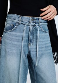 Světle modré džínové kalhoty s volným střihem, vysokým pasem, bočními kapsami, páskem na zavázání v pase a detaily s vybledlou texturou.