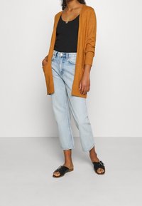 Dlouhý hořčicový cardigan z měkké pleteniny, spárovaný s černým topem bez rukávů, světle modrými džíny s vysokým pasem a černými skluzavkami s přezkou.