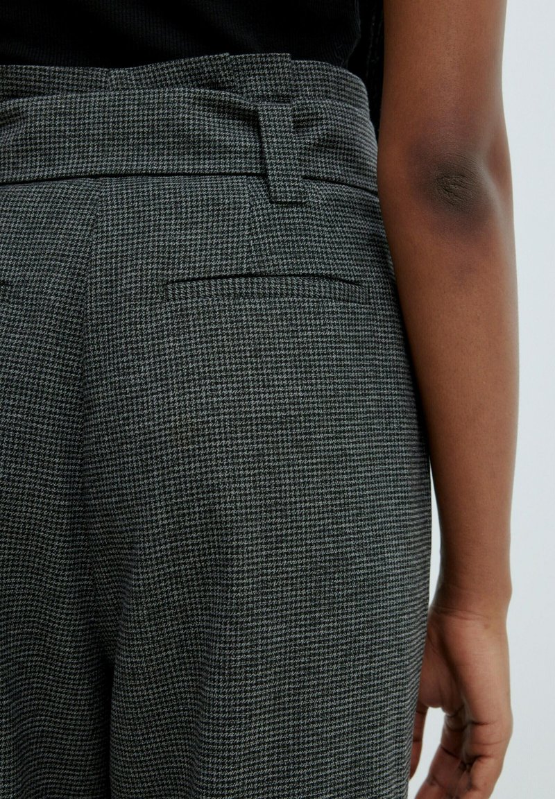 Nahaufnahme von karierten grauen Hosen mit Gürtelschlaufen und Seitentasche, getragen von einer Person mit dunkler Haut und sichtbarem Ellbogen.