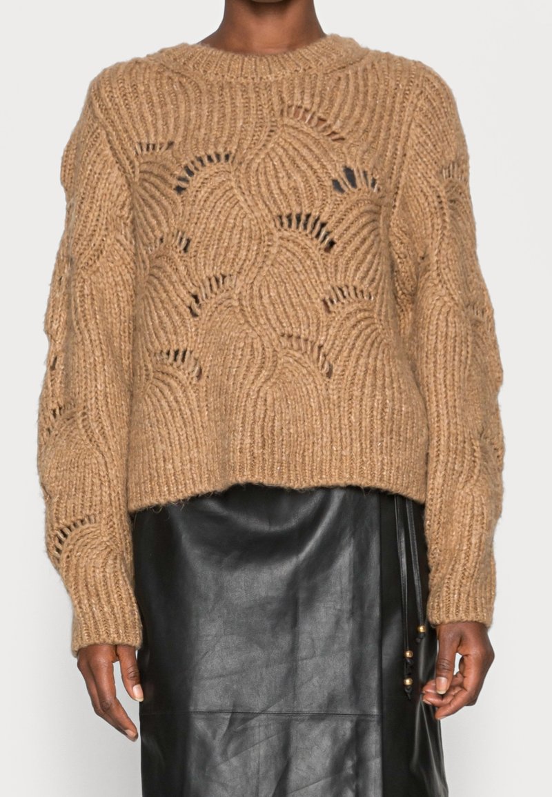 Pull en maille épaisse couleur beige avec motif ondulé, larges poignets côtelés et coupe décontractée, associé à une jupe en cuir noir.