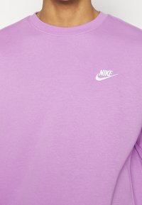 Fioletowa bluza Nike z miękkiej bawełny, z okrągłym dekoltem i białym haftowanym logo na lewej piersi. Gładka tekstura, luźny krój.